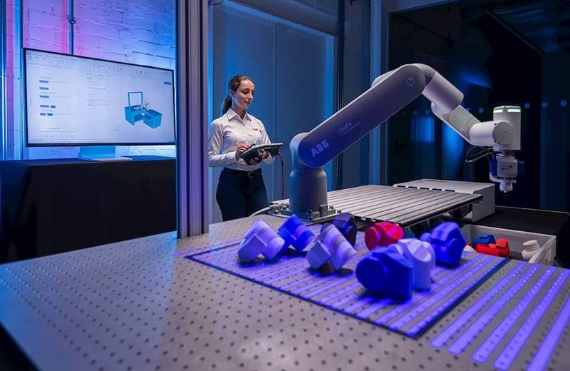 Mitarbeiterin bedient ein Tablet neben einem Roboterarm, der blaue Werkstücke auf einem Tisch aufnimmt (Quelle: The Robot Report)