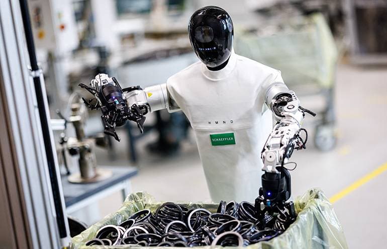 Humanoider Roboter greift mit beiden Händen Bauteile aus einem Behälter in einer Werkshalle