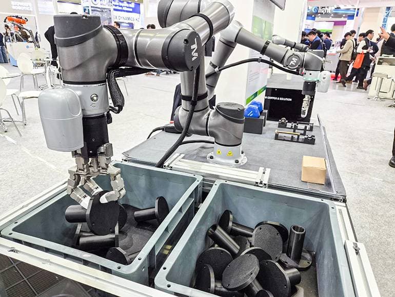 Roboterarm mit Mehrfinger-Greifer über Behältern mit Bauteilen (Symbolbild)